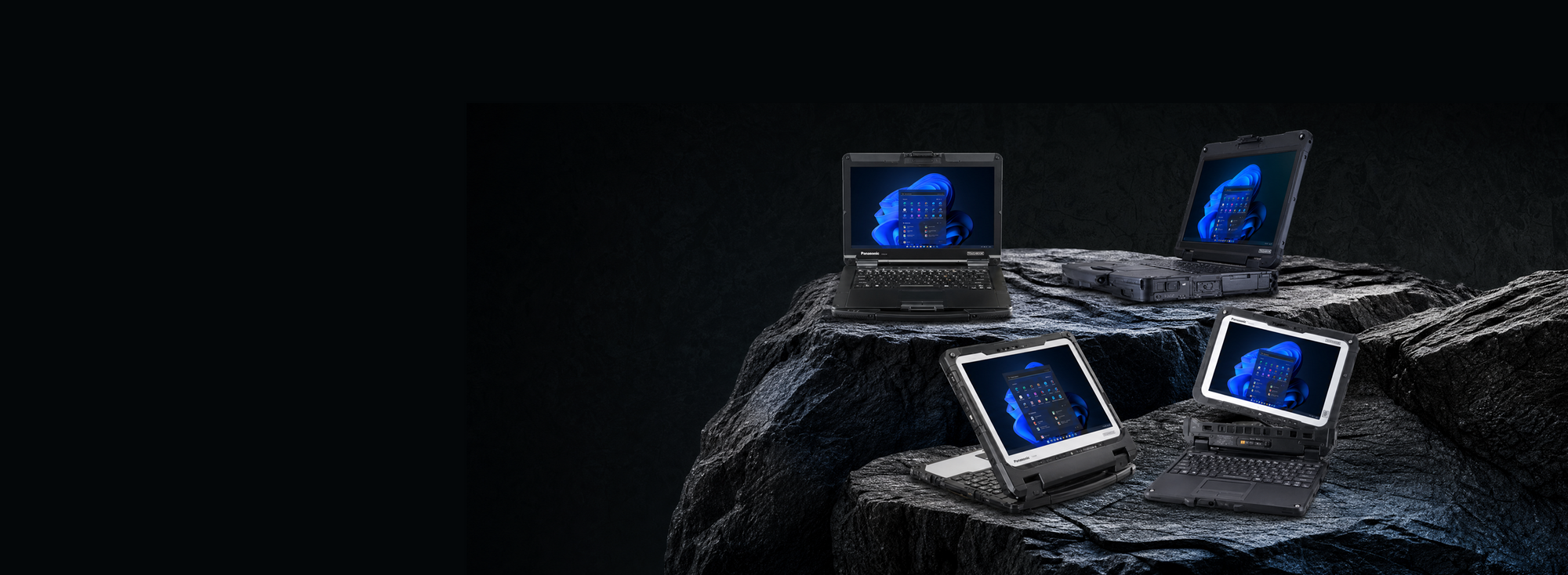 Four silver laptops with glowing blue screens are arranged on a dark rocky surface against a black background.