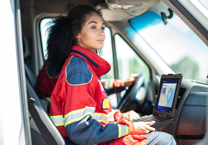 A woman in a red and navy high-visibility work jacket sits inside a vehicle, using a laptop on the dashboard. The jacket features yellow and silver reflective stripes.